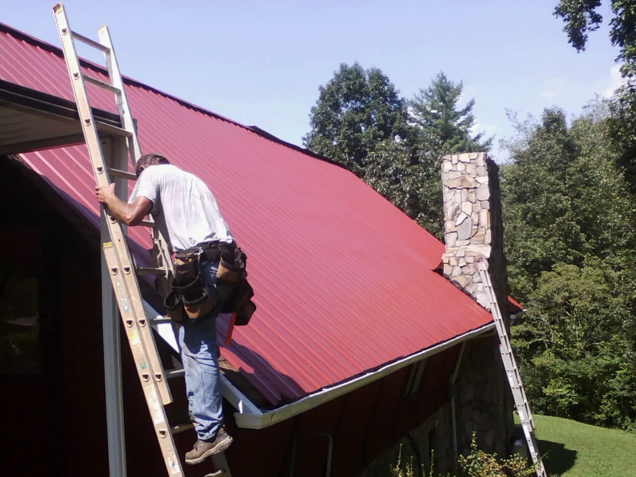 Roofer on a ladder beside a red metal roof and stone chimney — Soffit & Fascia Repair by Roofing Brothers in Minden